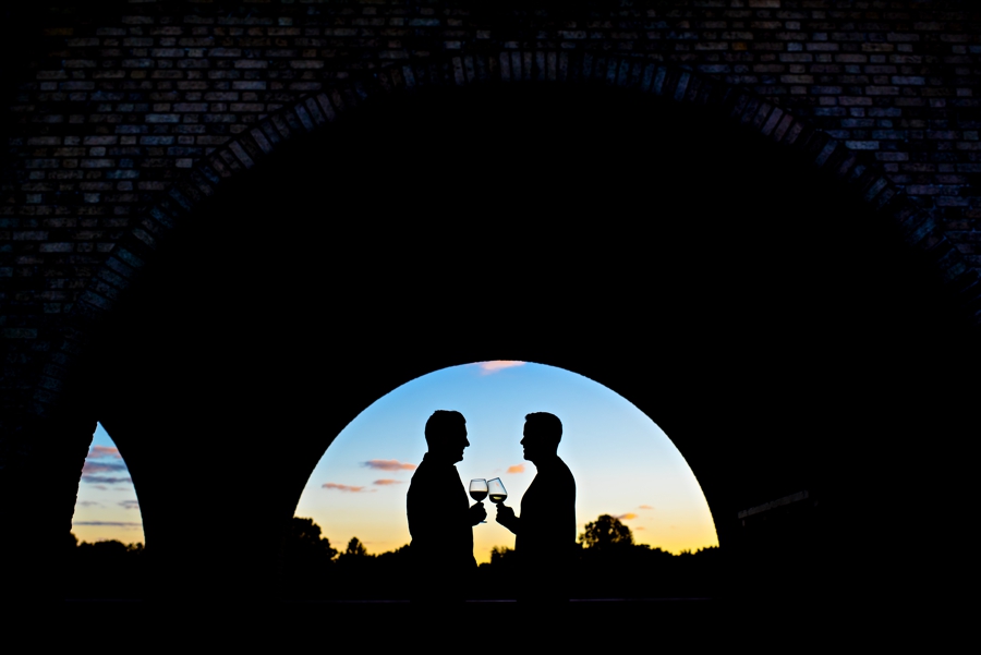 A silhouette of a gay couple cheersing glasses of wine with the setting sun behind them in their Philadelphia engagement session.